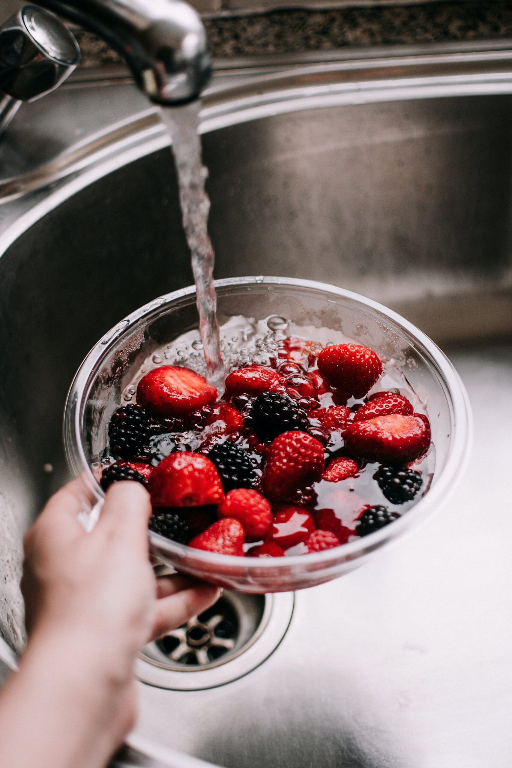 pexels-lisa-fotios-5137876 Hand holding a bowl of fresh strawberries and blackberries under running water in a kitchen sink, emphasizing fruit preparation and food hygiene.